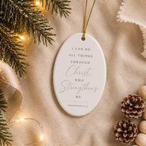 以下が含まれることがあります： 黒い文字で「I CAN DO ALL THINGS THROUGH Christ WHO Strengthens ME PHILIPPIANS 4:13」と印刷された楕円形の白いセラミック製オーナメント。オーナメントの上部には、吊り下げるための細い金色の紐が付いています。