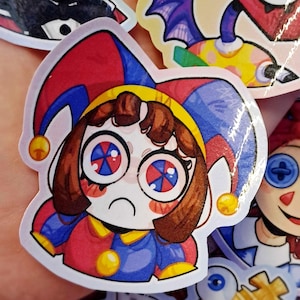 Pode incluir: Uma coleção de autocolantes de desenhos animados coloridos. O autocolante principal apresenta uma personagem de bobo da corte com uma roupa vermelha, azul e amarela, uma expressão triste e olhos grandes com círculos concêntricos vermelhos e azuis. Outros autocolantes mostram várias personagens de desenhos animados.