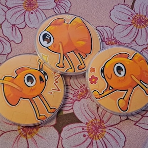 May include: Three round orange cartoon character buttons with black outlines and large eyes. The buttons are on a floral background with pink and white flowers. Each button features a different pose.