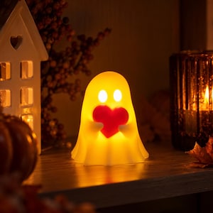 Könnte beinhalten: Eine kleine, leuchtende, geisterförmige Lampe, die ein warmes gelbes Licht ausstrahlt. Die Geisterfigur hat zwei ovale Augen und hält ein festes, leuchtend rotes Herz in der Mitte ihrer Brust. Die Lampe hat eine glatte, abgerundete und durchscheinende Oberfläche.
