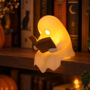 Könnte beinhalten: Eine kleine, weiße, geisterförmige Lampe, die ein warmes, sanftes Licht ausstrahlt. Die Geisterfigur sitzt und hält ein offenes, braunes Buch in den Händen. Die Augen bestehen aus zwei kreisförmigen, unbeleuchteten Aussparungen im Gesicht.