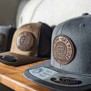 May include: Three baseball caps with flat brims are displayed on a wooden shelf. The caps are grey, brown, and dark grey, each featuring a circular leather patch with the text "BLAY PIPE GET PAID PIPELINER 2023".