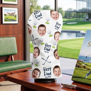 Puede incluir: Una funda blanca para la cabeza del palo de golf con un patr&oacute;n repetido de caras de ni&ntilde;os sonrientes, pelotas de golf y palos de golf cruzados. El texto "Best Dad by Par" tambi&eacute;n est&aacute; impreso en la funda. Un libro titulado "The Art of the Swing" es visible.