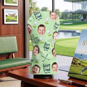 May include: A golf club head cover with a light green background, featuring multiple smiling faces, golf balls, and golf clubs. The text "Best Dad by Par" is repeated throughout the design. A book titled "The Art of the Swing" is also visible.