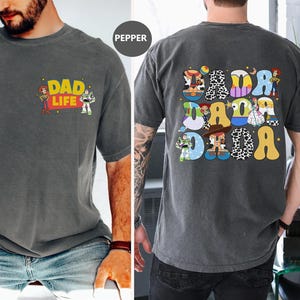 May include: A gray t-shirt with a distressed look featuring a cartoon image of Woody and Buzz Lightyear from Toy Story. The text "Dad Life" is printed on the shirt. The back of the shirt features a colorful design with the word "Dada" spelled out in a retro style with different Toy Story characters.