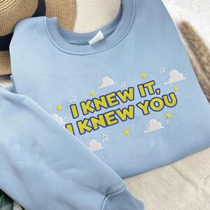 May include: A light blue crewneck sweatshirt featuring embroidered text that reads I KNEW IT, I KNEW YOU in yellow with a dark blue outline. The design includes small yellow stars, white clouds, and musical note symbols scattered around the text.