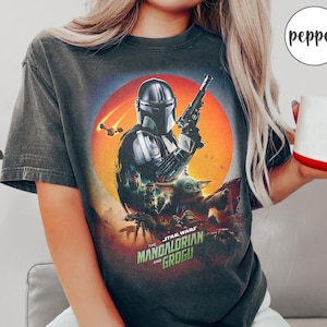 Op de afbeelding: Donkergrijs t-shirt met een afbeelding uit de serie "The Mandalorian". Het ontwerp toont de Mandalorian, Grogu en de tekst "The Star Wars Mandalorian and Grogu" tegen een oranje en rode achtergrond. Een casual kledingstuk.