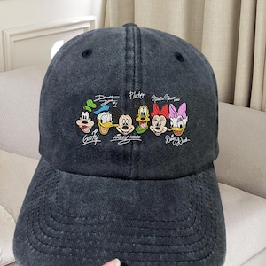 May include: A dark grey baseball cap with embroidered Disney characters, including Mickey Mouse, Donald Duck, and Goofy. The cap has a curved peak and the characters' names are written above them.