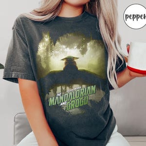 May include: Dark gray t-shirt featuring a graphic of Grogu from The Mandalorian series. The image shows Grogu in a forest setting, with the text "Star Wars The Mandalorian and Grogu" in green and white.