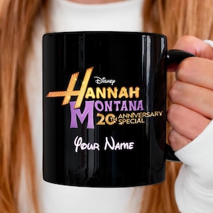 Puede incluir: Taza de cer&aacute;mica negra con el logotipo de Hannah Montana 20th Anniversary Special en dorado, morado y blanco. La taza incluye el logotipo de Disney y espacio para un nombre personalizado en escritura blanca. Una taza de caf&eacute; cl&aacute;sica.