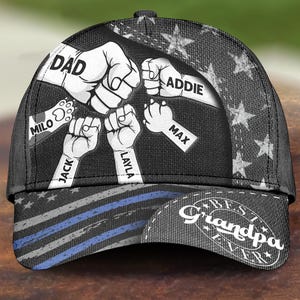 May include: Black baseball cap with a design featuring fists, stars, and stripes. The cap has the words "DAD", "ADDIE", "MAX", "JACK", "LAYLA", and "MILO" on it. The text "Best Grandpa Ever" is also visible.