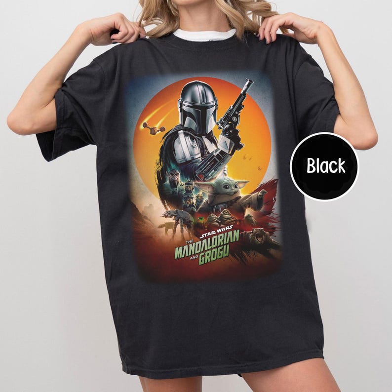 Op de afbeelding: Zwart t-shirt met een afbeelding van "The Mandalorian and Grogu". Het ontwerp toont de Mandalorian, Grogu en andere personages tegen een zonsondergang. De tekst "The Mandalorian and Grogu" staat onderaan.