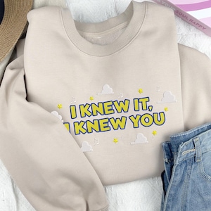 May include: A beige crewneck sweatshirt featuring embroidered text that reads I KNEW IT, I KNEW YOU in yellow with blue outlines. The design includes small white embroidered clouds, yellow stars, and musical note symbols scattered around the lettering.