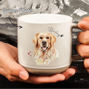 May include: Cream-colored ceramic mug featuring a watercolor portrait of a Golden Retriever. The mug has the name "Bailey" and paw print, with the text "Custom Photo" and small red hearts. The mug is held by a person.