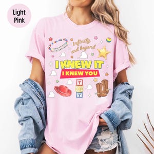 May include: A light pink t-shirt featuring a graphic print with the text "infinity and beyond", "I KNEW IT", and "I KNEW YOU". The design includes illustrations of a beaded bracelet, a yellow star, a red cowboy hat, toy blocks, and boots.