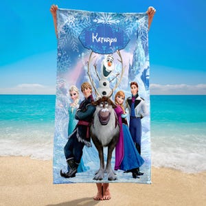 May include: A rectangular beach towel featuring characters from the film Frozen, including Elsa, Anna, Kristoff, Olaf, and Sven. The towel has a blue background with snowflakes and the name "Kathryn" in a decorative banner. The towel is held up on a beach.