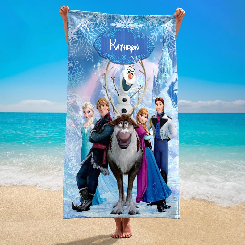 May include: A rectangular beach towel featuring characters from the film Frozen, including Elsa, Anna, Kristoff, Olaf, and Sven. The towel has a blue background with snowflakes and the name "Kathryn" in a decorative banner. The towel is held up on a beach.