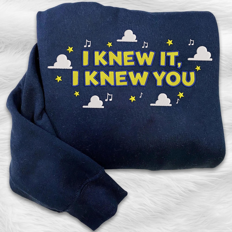 May include: A navy blue sweatshirt featuring embroidered text that reads "I KNEW IT, I KNEW YOU" in yellow with a blue outline. The design is accented with white clouds, yellow stars, and small musical note symbols scattered around the lettering.