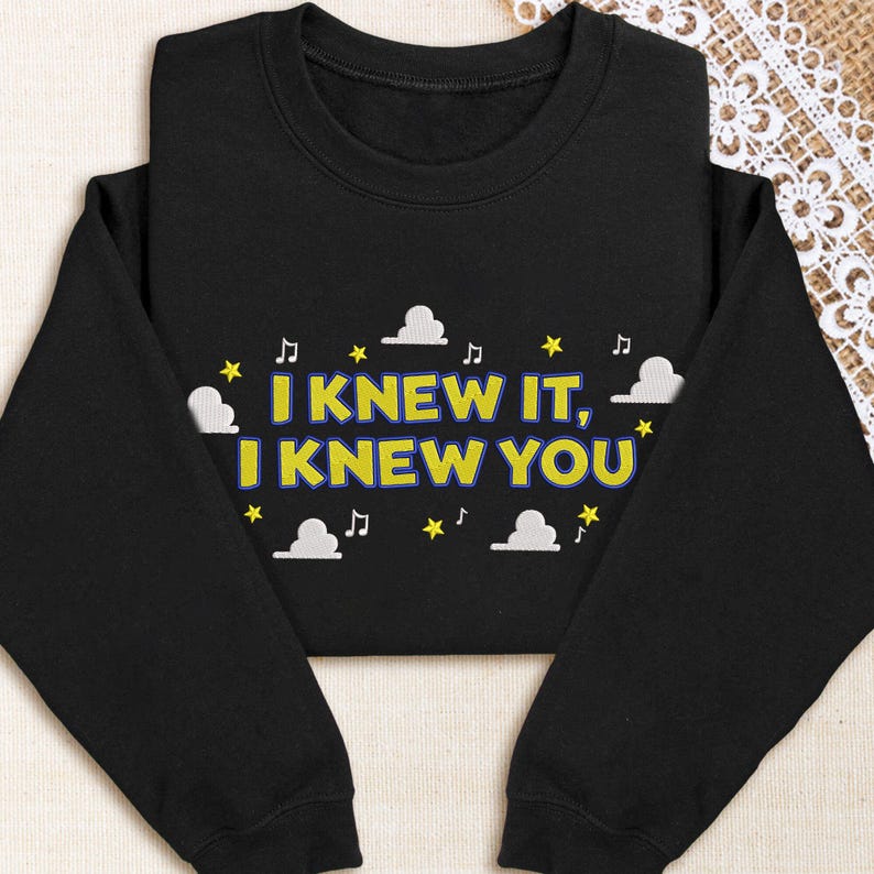 May include: A black long-sleeve sweatshirt featuring the embroidered yellow text "I KNEW IT, I KNEW YOU" with a blue outline. The design is surrounded by white embroidered clouds, yellow stars, and small black musical note symbols.