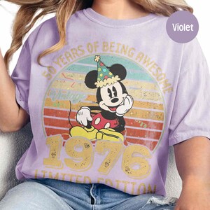 Puede incluir: Camiseta violeta con un dise&ntilde;o de Mickey Mouse con un gorro de fiesta. El dise&ntilde;o incluye el texto "50 YEARS OF BEING AWESOME 1976 LIMITED EDITION" en letras doradas, sobre un fondo retro de puesta de sol. Una camiseta divertida y festiva.