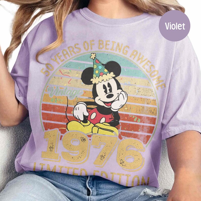 Puede incluir: Camiseta violeta con un dise&ntilde;o de Mickey Mouse con un gorro de fiesta. El dise&ntilde;o incluye el texto "50 YEARS OF BEING AWESOME 1976 LIMITED EDITION" en letras doradas, sobre un fondo retro de puesta de sol. Una camiseta divertida y festiva.