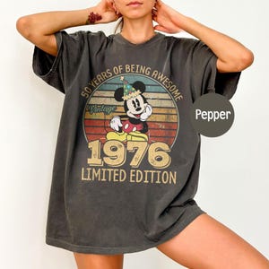 Puede incluir: Camiseta color pimienta con un dise&ntilde;o vintage de Mickey Mouse con gorro de fiesta. El gr&aacute;fico incluye el texto "50 Years of Being Awesome", "Vintage", "1976" y "Limited Edition". La camiseta tiene un corte holgado y mangas cortas.