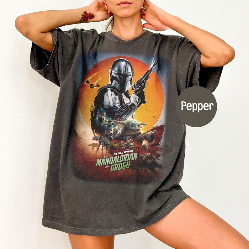 Op de afbeelding: Donkergrijs t-shirt met een afbeelding van "The Mandalorian and Grogu". Het ontwerp toont de Mandalorian met een blaster, Grogu en andere personages tegen een zonsondergang. Het shirt heeft een relaxte pasvorm.