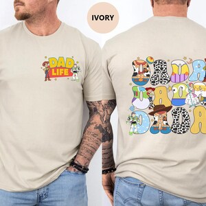 May include: A beige t-shirt with a graphic of Toy Story characters spelling out "DAD DAD DAD" on the back. The front of the shirt has a graphic of Woody and Buzz Lightyear with the text "DAD LIFE".