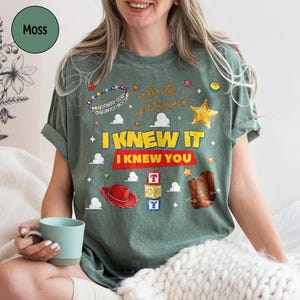 May include: A moss green short-sleeve t-shirt featuring a graphic print with the text "I KNEW IT I KNEW YOU" and "infinity and beyond". The design includes illustrations of a red cowboy hat, brown boots, alphabet blocks, a star, and beads.