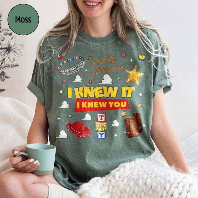 May include: A moss green short-sleeve t-shirt featuring a graphic print with the text "I KNEW IT I KNEW YOU" and "infinity and beyond". The design includes illustrations of a red cowboy hat, brown boots, alphabet blocks, a star, and beads.