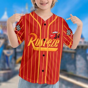 May include: Red and yellow pinstripe baseball jersey with the word "Rusteze" in yellow script across the chest. A cartoon car graphic is on the right chest. Checkered flags are on the sleeves. The jersey has black buttons.