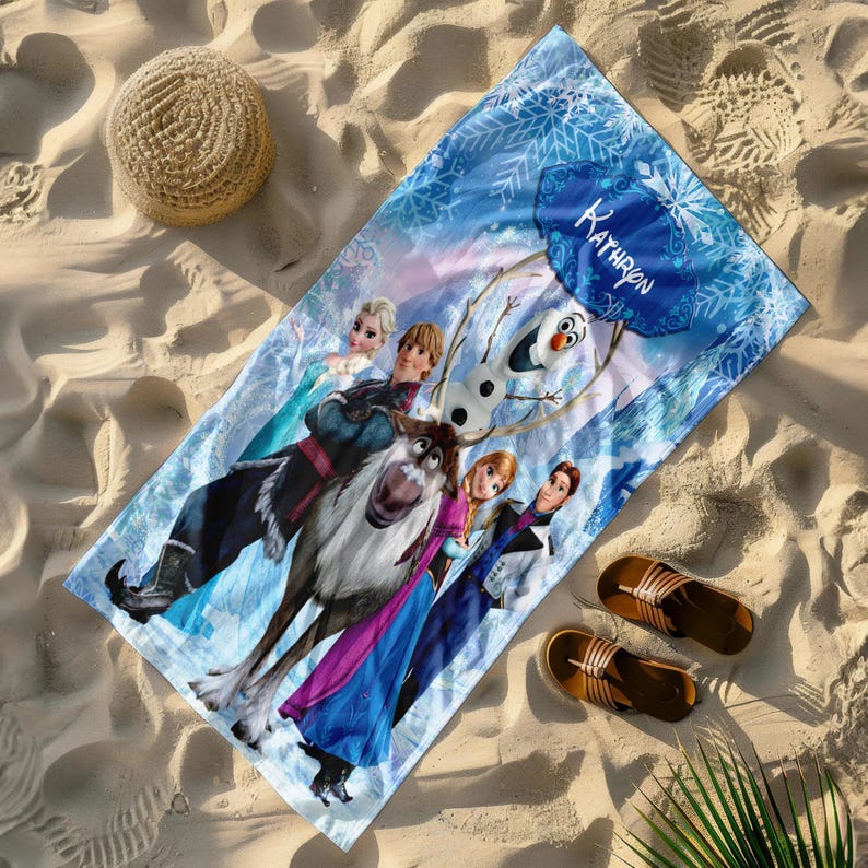 May include: A blue beach towel featuring characters from the film Frozen, including Elsa, Anna, Kristoff, Olaf, and Sven. The towel has a personalized name, "Kathron," in a blue snowflake design. A straw hat and brown sandals are also visible.