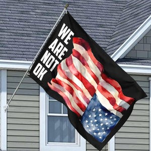 May include: A black flag with the words "We Are Not Ok" in white text. The flag features a watercolor design of the American flag.