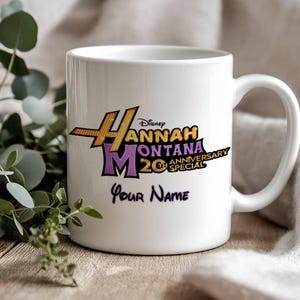 Puede incluir: Taza de cer&aacute;mica blanca con el logo "Hannah Montana 20th Anniversary Special" en naranja, morado y negro. La taza tambi&eacute;n incluye el texto "Your Name" en una fuente cursiva. Un regalo perfecto para los fans.