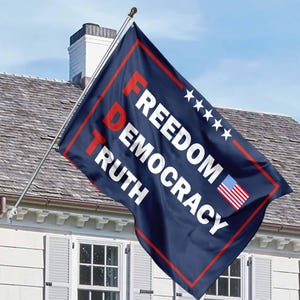 May include: A blue flag with white text that reads "FREEDOM DEMOCRACY TRUTH" and four white stars. The flag also has a small American flag in the bottom right corner.
