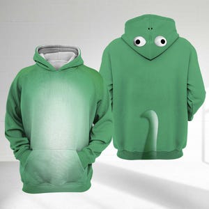 May include: Green hooded sweatshirt with a kangaroo pocket and a white-lined hood. The back of the hoodie features a frog design with large, googly eyes on the hood and a white tail. The sweatshirt is made of a soft material.