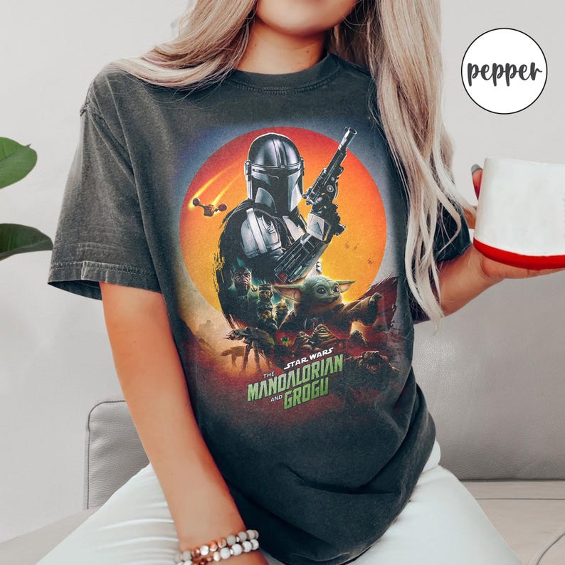 Op de afbeelding: Donkergrijs t-shirt met een afbeelding uit de serie "The Mandalorian". Het ontwerp toont de Mandalorian, Grogu en de tekst "The Star Wars Mandalorian and Grogu" tegen een oranje en rode achtergrond. Een casual kledingstuk.