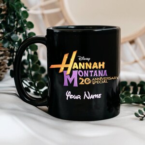 Puede incluir: Taza de cer&aacute;mica negra con el texto "Hannah Montana 20th Anniversary Special" en dorado, morado y blanco. La taza tambi&eacute;n incluye el logotipo de Disney y espacio para un nombre personalizado.