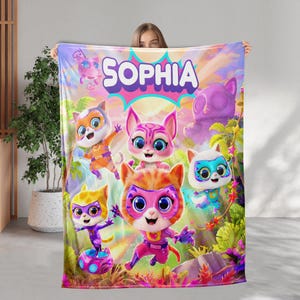 May include: A colorful blanket featuring cartoon cat characters with superhero masks. The blanket has the name "SOPHIA" in large, bubble letters. The design includes vibrant colors like pink, orange, blue, and purple, creating a playful and whimsical aesthetic.
