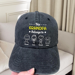 May include: A dark gray baseball cap with the text "This Grandpa Belongs to" in white and yellow. Below the text are stick figure illustrations of children and a dog, with names below each figure.