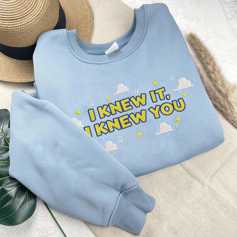 May include: A light blue crewneck sweatshirt featuring embroidered text that reads I KNEW IT, I KNEW YOU in yellow with a dark blue outline. The design includes small yellow stars, white clouds, and musical note symbols scattered around the text.