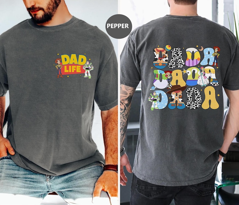 May include: A gray t-shirt with a distressed look featuring a cartoon image of Woody and Buzz Lightyear from Toy Story. The text "Dad Life" is printed on the shirt. The back of the shirt features a colorful design with the word "Dada" spelled out in a retro style with different Toy Story characters.