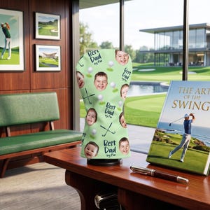 Puede incluir: Una funda para la cabeza del palo de golf de color verde claro con caras sonrientes, pelotas de golf y palos de golf. El texto "Best Dad by Par" se repite. Tambi&eacute;n se ve un libro titulado "The Art of the Swing".
