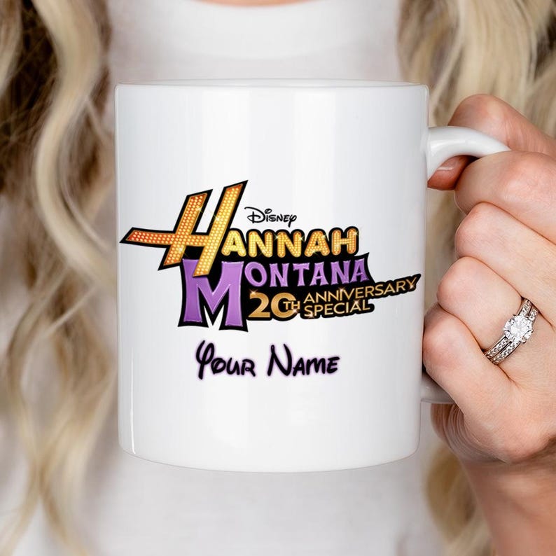 Puede incluir: Taza de cer&aacute;mica blanca con el logotipo de Hannah Montana 20th Anniversary Special en dorado, morado y negro. La taza tambi&eacute;n incluye el texto "Your Name" en una fuente de escritura. La taza est&aacute; sostenida por una persona.