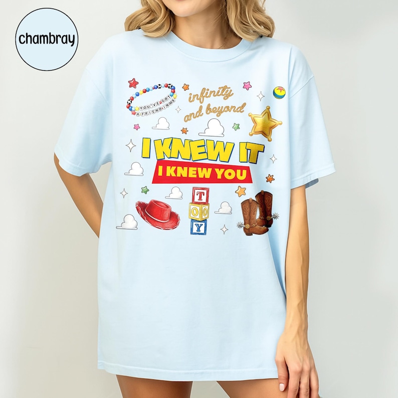 May include: A light blue t-shirt featuring a graphic print with the text "I KNEW IT I KNEW YOU" and "infinity and beyond". The design includes a beaded bracelet, a red cowboy hat, brown cowboy boots, a gold star, and toy blocks spelling TOY.