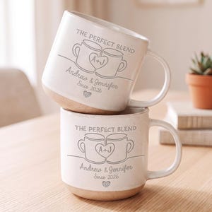 Peut inclure: Deux tasses en céramique blanc cassé avec une finition mouchetée et une base beige. Chaque tasse présente le texte THE PERFECT BLEND, un graphique de deux tasses reliées par un cœur avec A+J, les noms Andrew & Jennifer, Since 2026 et un cœur.