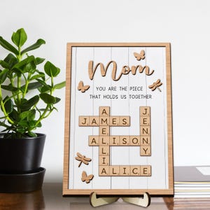 May include: A wooden sign with the word "Mom" in cursive script and the text "You are the piece that holds us together". The sign features a scrabble tile crossword puzzle with the names James, Alison, Jenny and Alice.