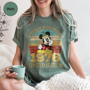 Puede incluir: Camiseta verde musgo con un dise&ntilde;o vintage de Mickey Mouse celebrando su 50 aniversario. El dise&ntilde;o incluye el texto "50 Years of Being Awesome" y "1976 Limited Edition" en dorado. La camiseta es de estilo casual.