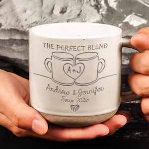 May include: A white ceramic mug with a speckled finish featuring an engraved design of two mugs joined by a heart containing the letters A+J. The text reads THE PERFECT BLEND, Andrew & Jennifer, and Since 2026, with a small heart icon below.