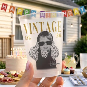 May include: A clear plastic party cup featuring gold text that reads VINTAGE EST. 1986. The cup displays a black and white photograph of a child wearing sunglasses, with the name Helena printed in a black script font below the image.